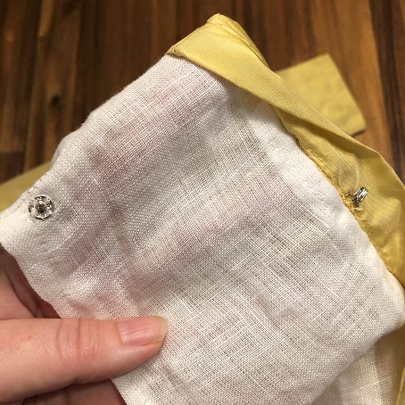 A close up of a silver snap fastener attached to white linen on a sleeve, and a silver hook attached to the inside seam of a gold silk sleeve