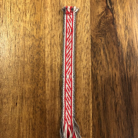 A close up of a piece of red, light grey, and dark grey tablet woven trim with a pattern that looks like angled box shapes